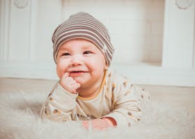 A smiling baby wearing a striped beanie and cozy outfit lies on a soft carpet indoors.